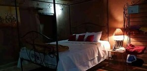 Een eenpersoonsbed met metalen frame, een nachtkastje met lamp en een wandplank met een rood voorwerp.