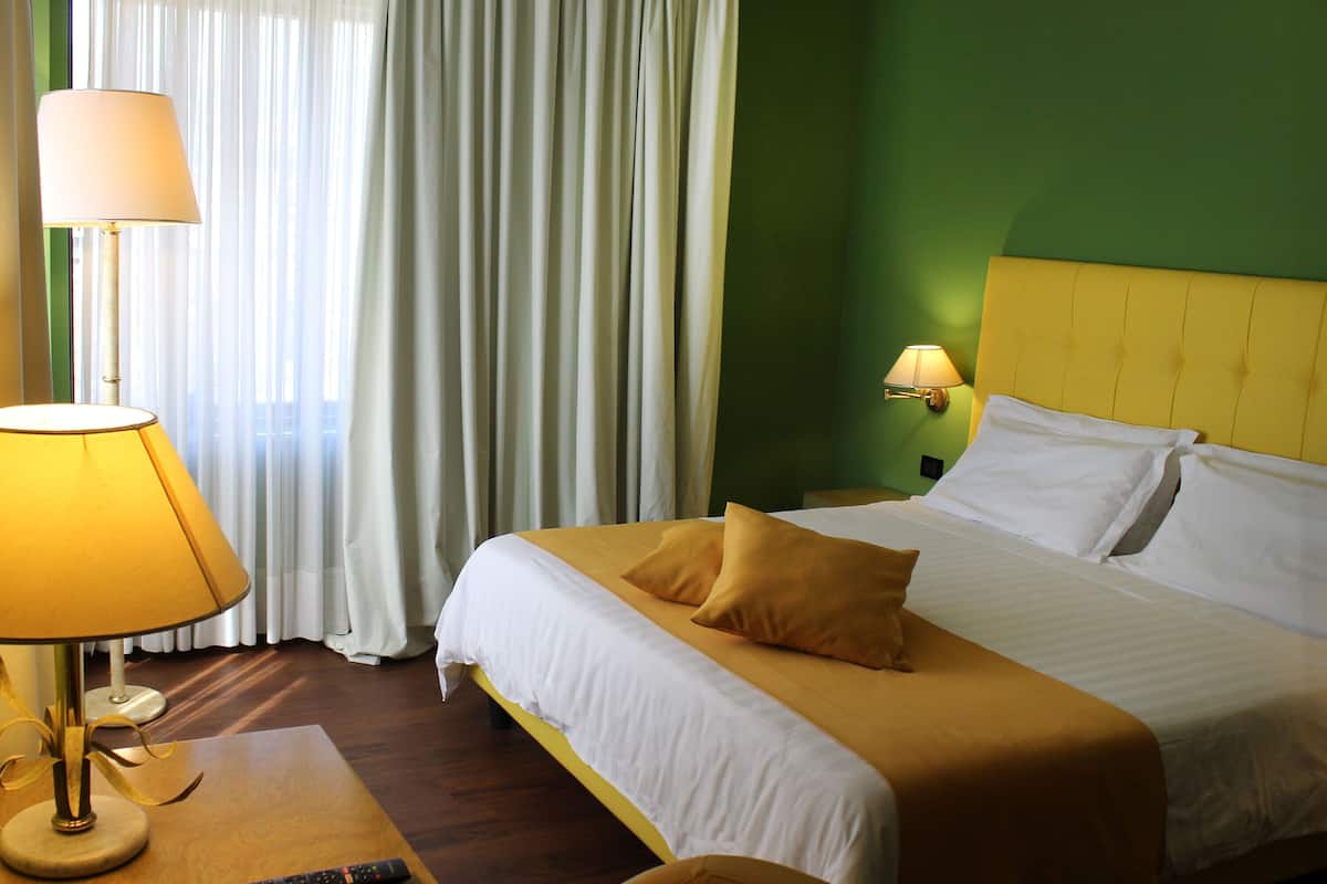 Una habitación de hotel con cama, mesita de noche, lámpara y ventana con cortinas.