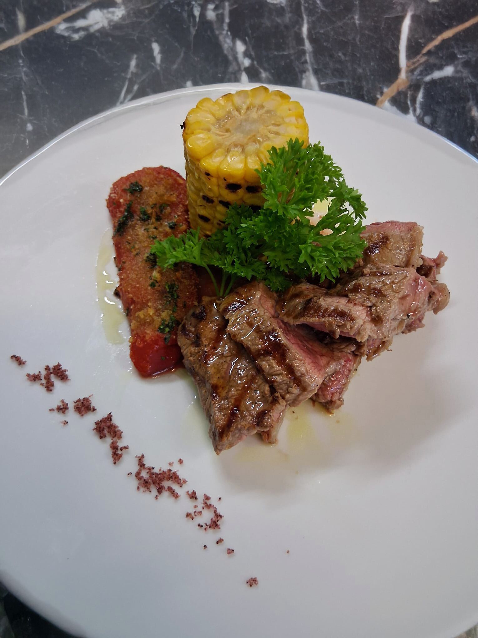 A plate with grilled steak, a grilled corn on the cob, and a grilled vegetable, possibly a potato, garnished with parsley.
