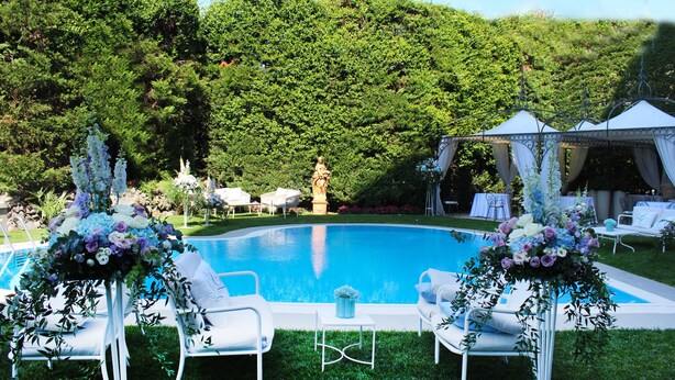 Un área junto a la piscina con sillones blancos, una carpa y arreglos florales.