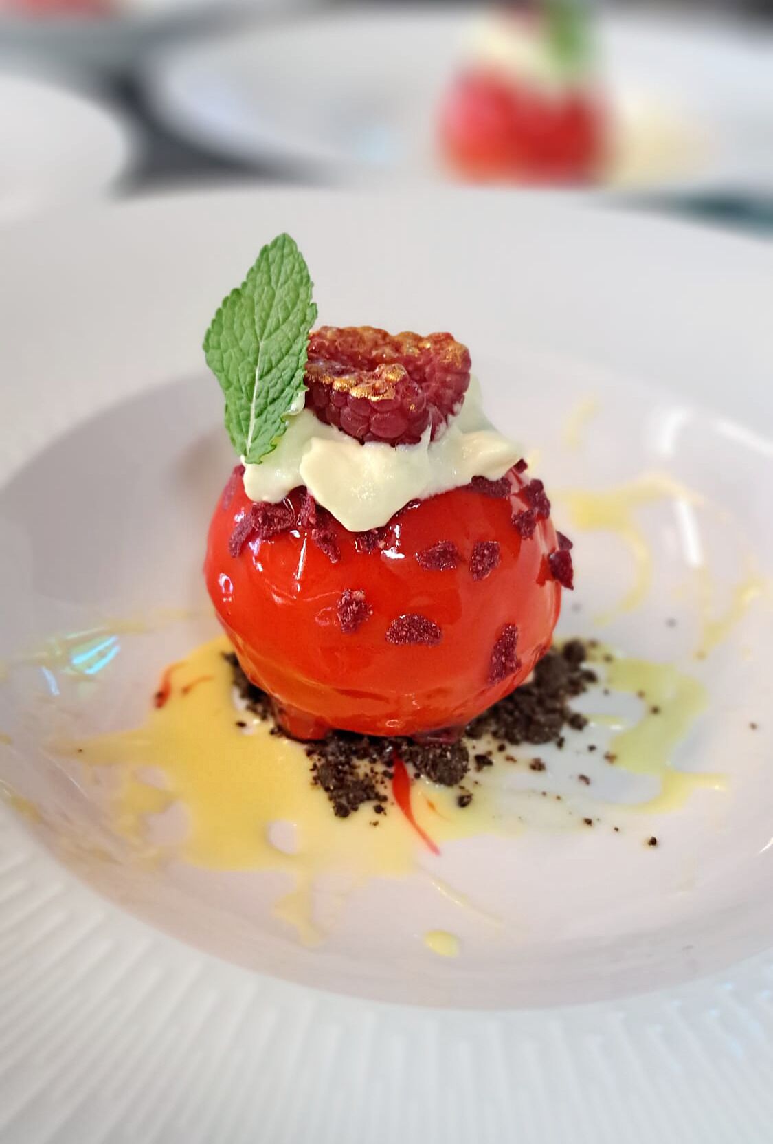 A dessert on a white plate with a mint leaf garnish, raspberry, and a creamy topping.