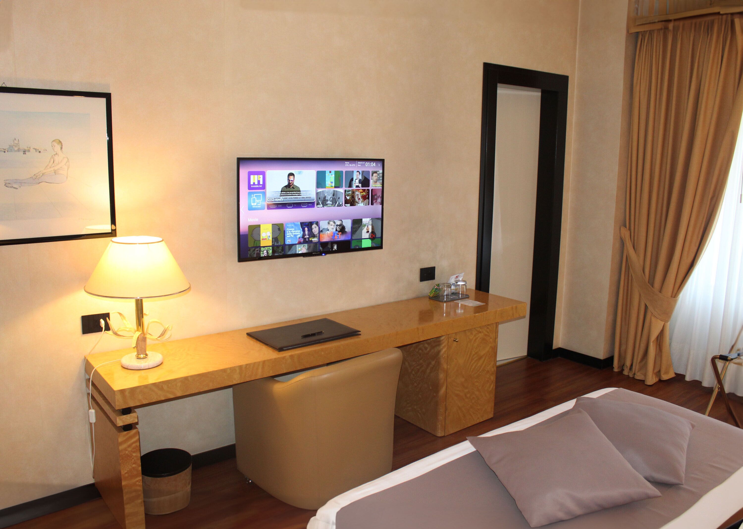 Una habitación de hotel con cama, escritorio con computadora portátil, televisión mostrando una interfaz de videollamada, lámpara y un cuadro en la pared.