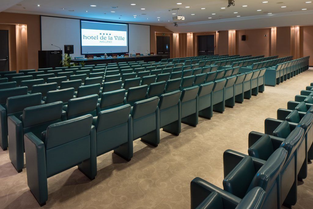 A conference room with rows of teal chairs, a large screen displaying 'hotel de la Ville AVELLINO', and a podium with a laptop.