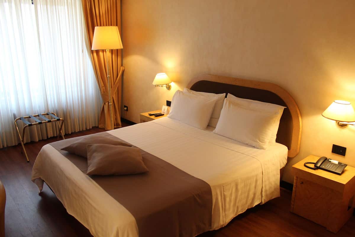 Habitación de hotel con una cama grande, dos mesitas de noche con lámparas, una silla y un ventanal con cortinas.