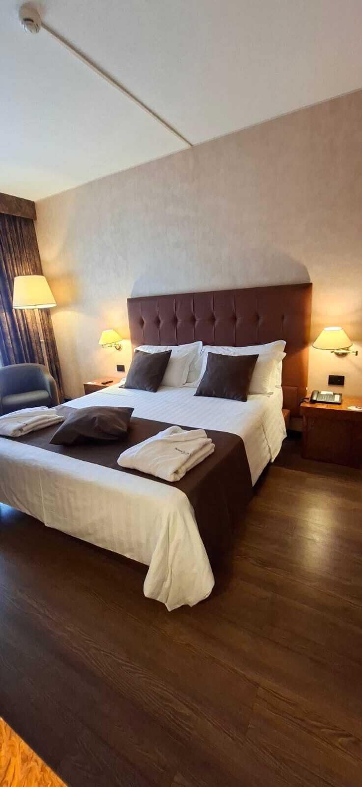 Quarto de hotel com uma cama grande, duas luminárias de cabeceira, uma cadeira e piso de madeira.