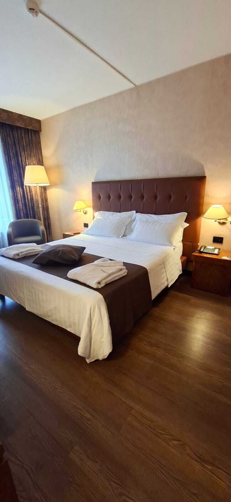 A hotel room with a large bed, two bedside lamps, a chair, and a wooden floor.