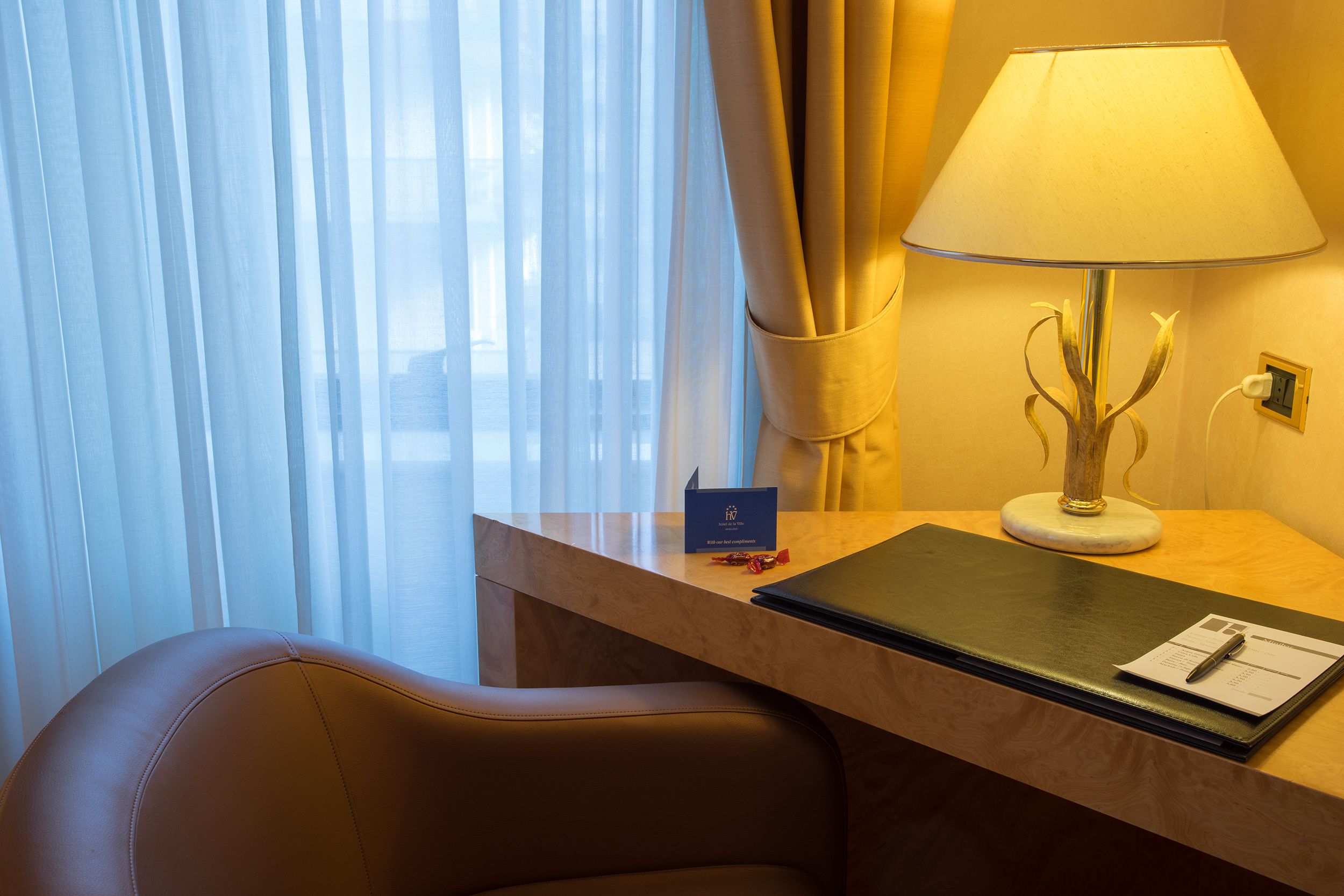 A hotel room with a desk, a lamp, a chair, and a view through sheer curtains.