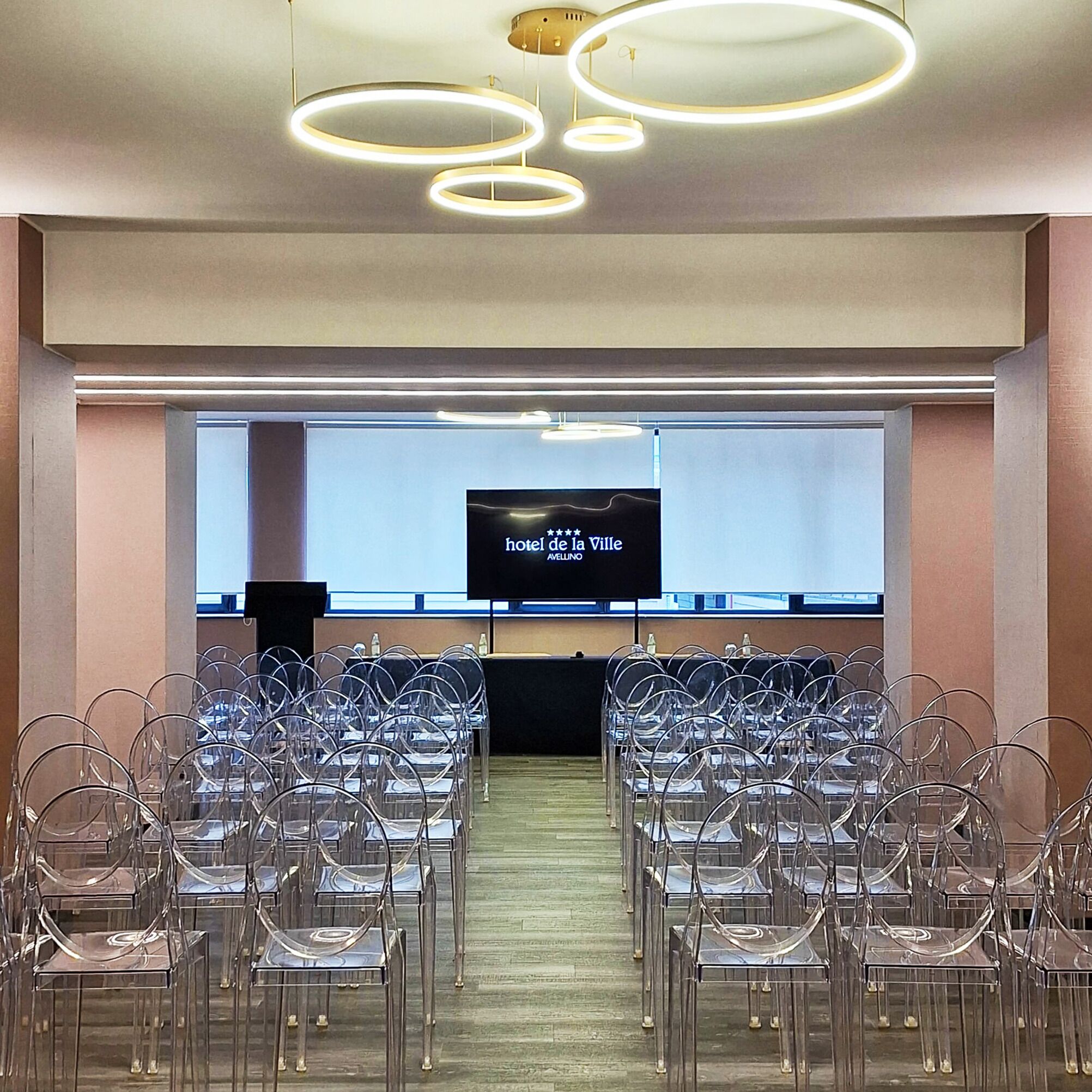 A conference room with a large screen displaying hotel information, clear plastic chairs arranged in rows, and modern circular ceiling lights.