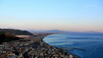 A coastal cityscape with dense urban development along the waterfront, featuring a mix of high-rise and low-rise buildings, a prominent road running parallel to the coast, and a clear view of the sea and distant mountains.