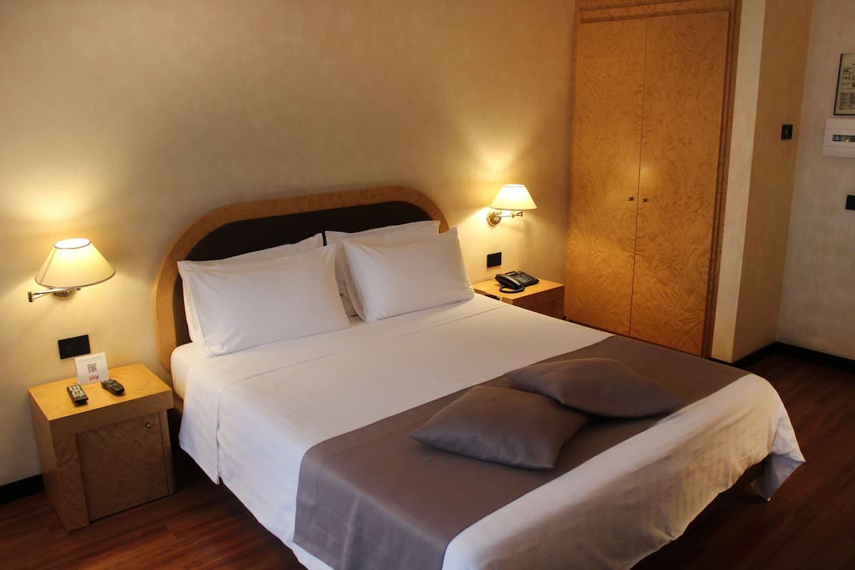 Una cama bien hecha con ropa de cama blanca y gris, dos lámparas de noche, una mesita de noche con un control remoto y un armario de madera.
