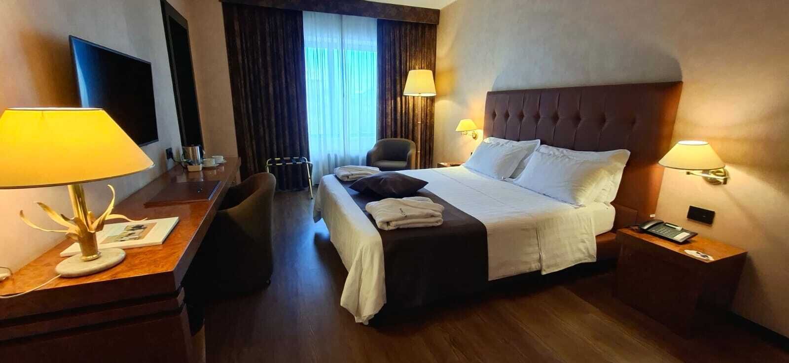 Habitación de hotel con cama, lámparas de noche, un escritorio con lámpara, una silla y vistas al exterior por la ventana.
