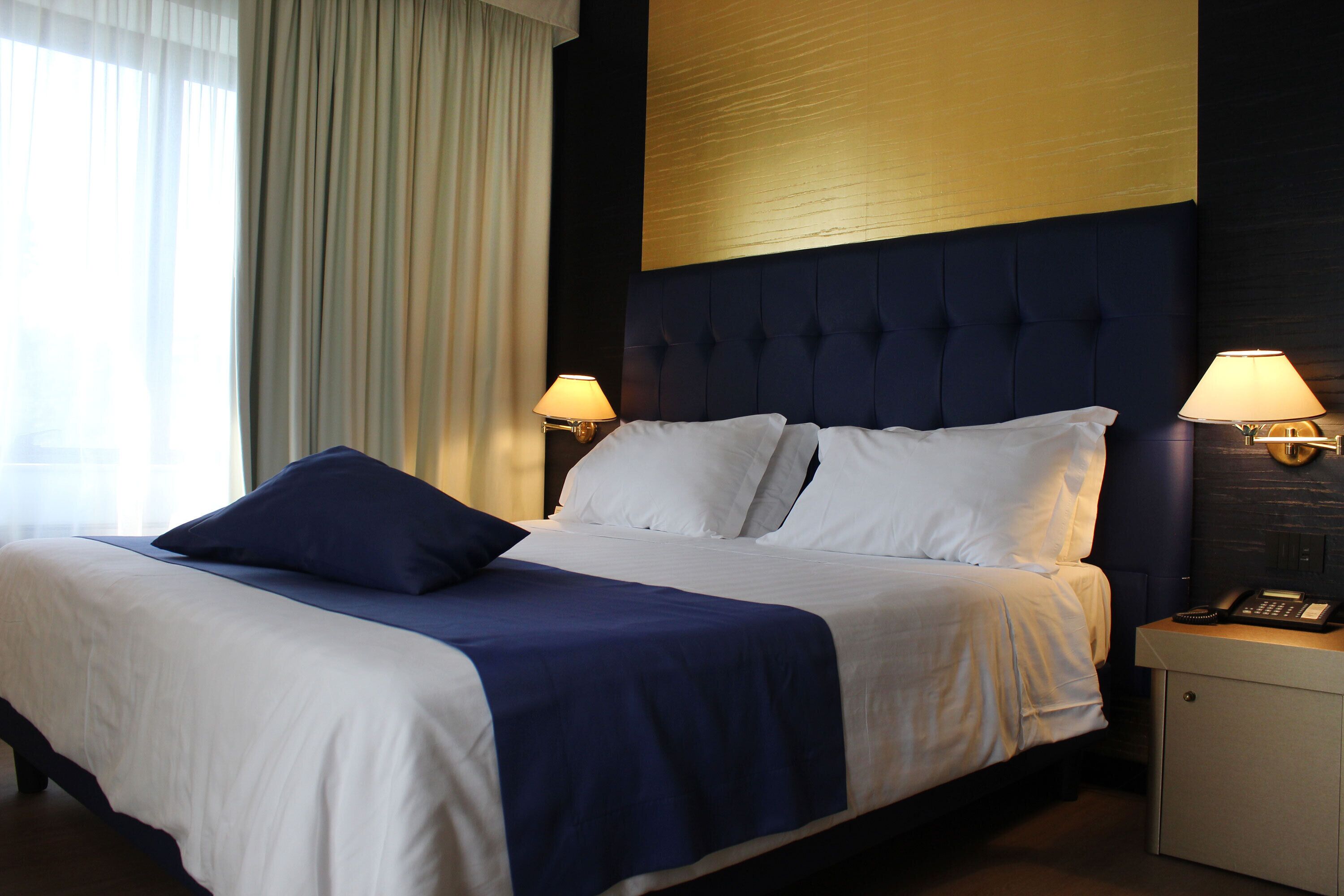 A hotel room with a large bed, two bedside lamps, a nightstand with a phone, and a view of the outdoors through a window with curtains.