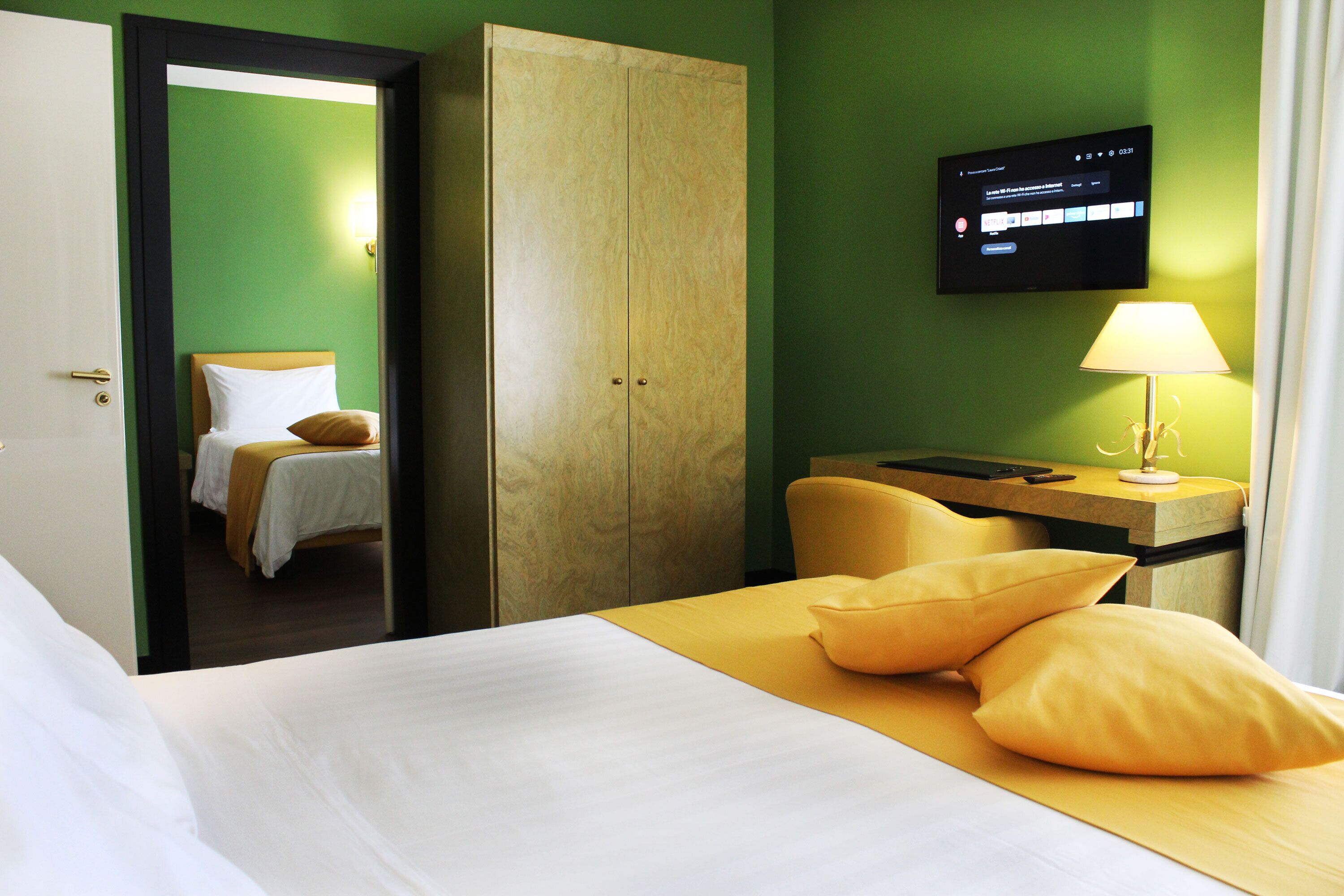 A hotel room with a bed, a wardrobe, a desk with a lamp, and a television on the wall.