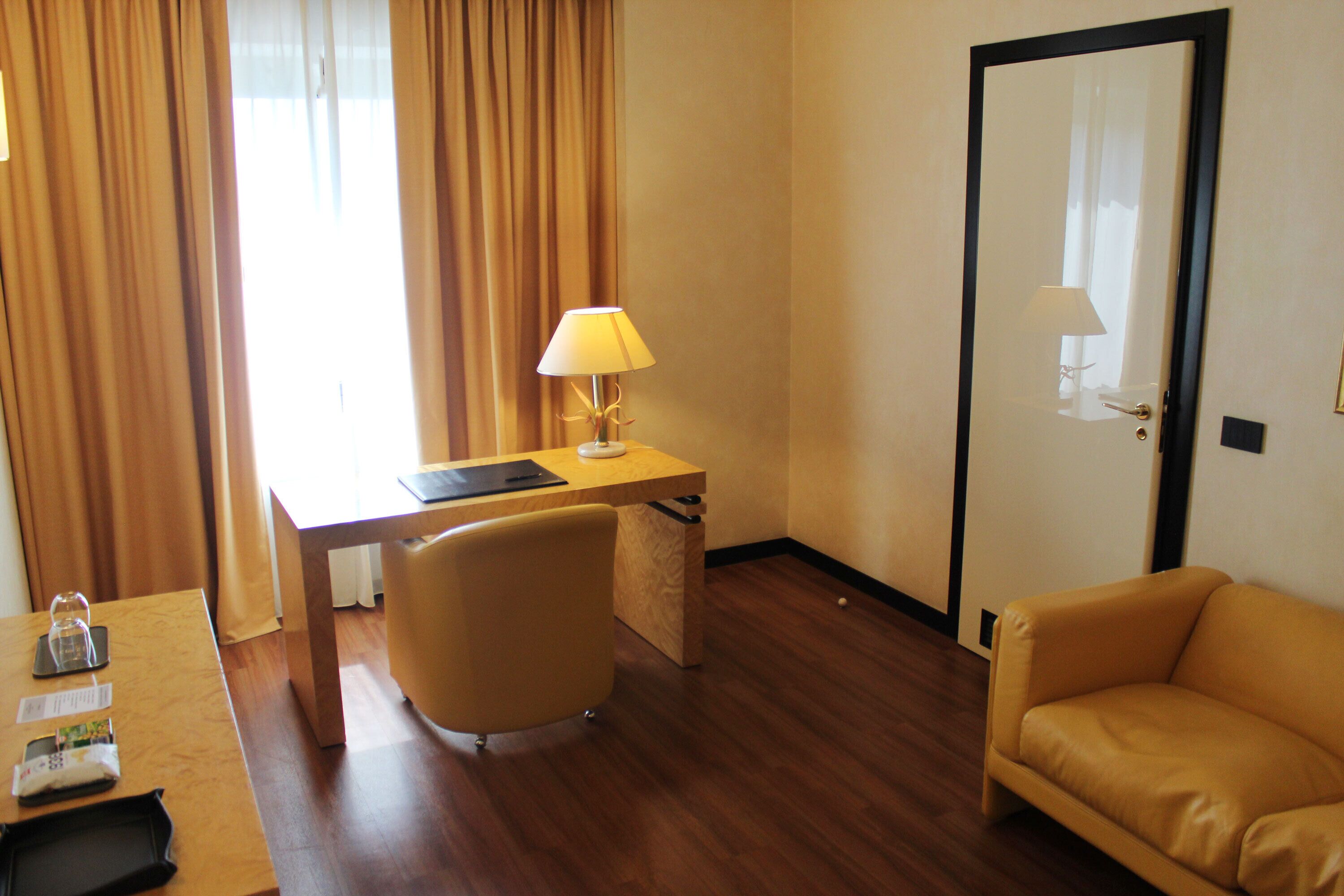A hotel room with a wooden desk, a chair, a sofa, and a lamp.