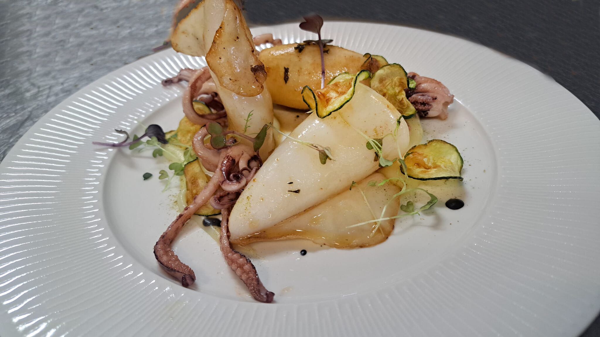 A plate of food with octopus, zucchini, and possibly potatoes, garnished with microgreens.
