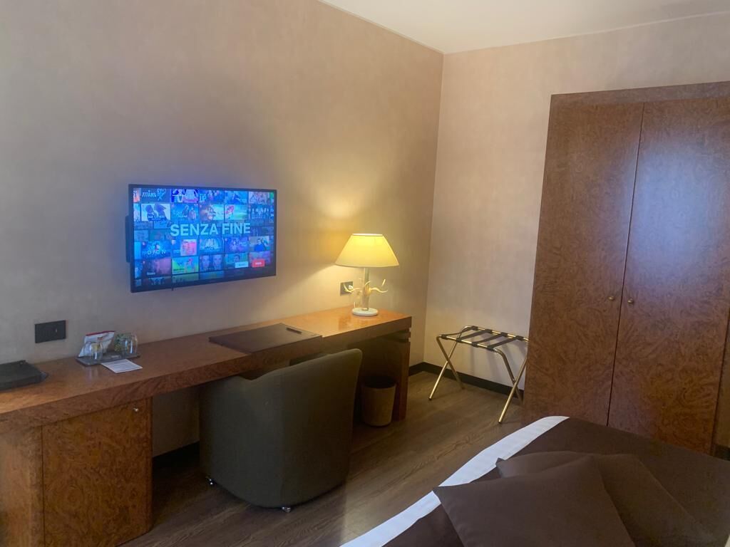 A hotel room with a wooden desk, a television mounted on the wall, a lamp, a chair, and a wardrobe.