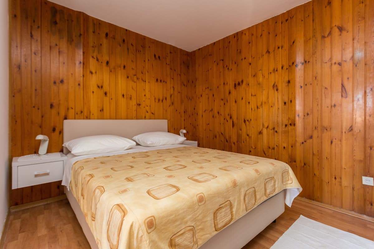A bedroom with wooden paneling, a bed with a patterned bedspread, a nightstand with a lamp, and a white rug.