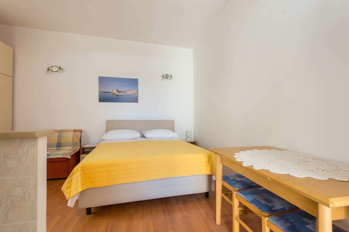 A bedroom with a bed, a bedside table with a lace tablecloth, a chair, and a wall-mounted picture.