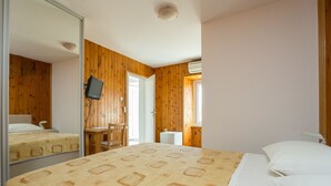A bedroom with a wooden paneled wall, a bed with a patterned bedspread, a desk with a chair, and a television mounted on the wall. - Guest House Luce (Dubrovnik)