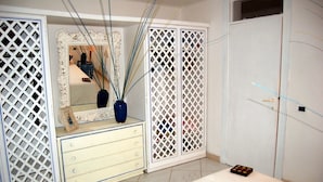 Un dormitorio con un armario blanco, un aparador con cajones, un espejo con marco decorativo y un florero con tallos altos.