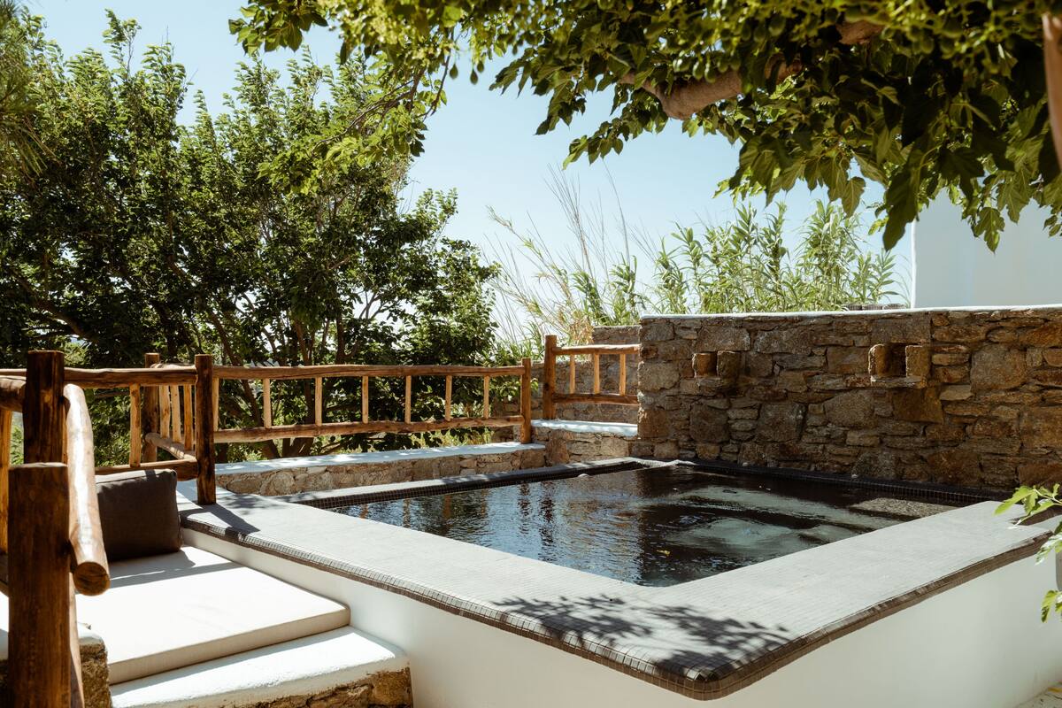 A stone-walled hot tub with a wooden deck and steps leading to it, surrounded by lush greenery.