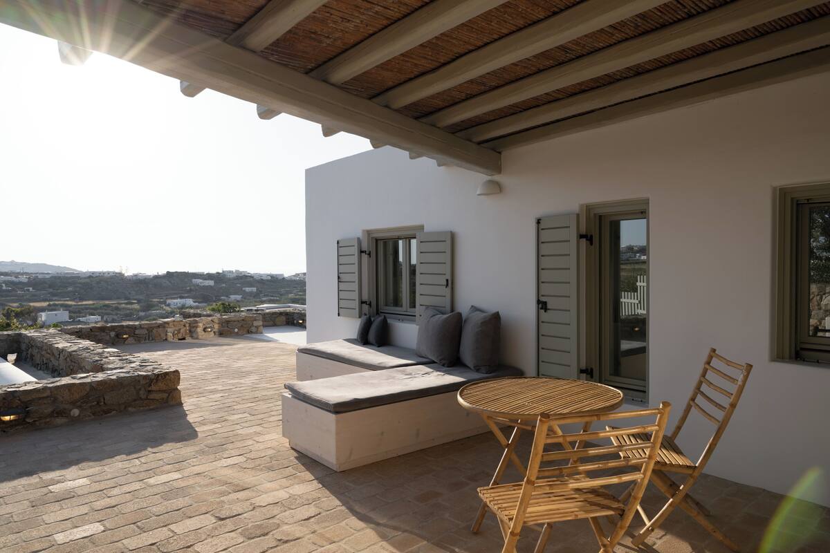 A terrace with a stone floor, a wooden table with chairs, a cushioned seating area, and a view of the landscape.