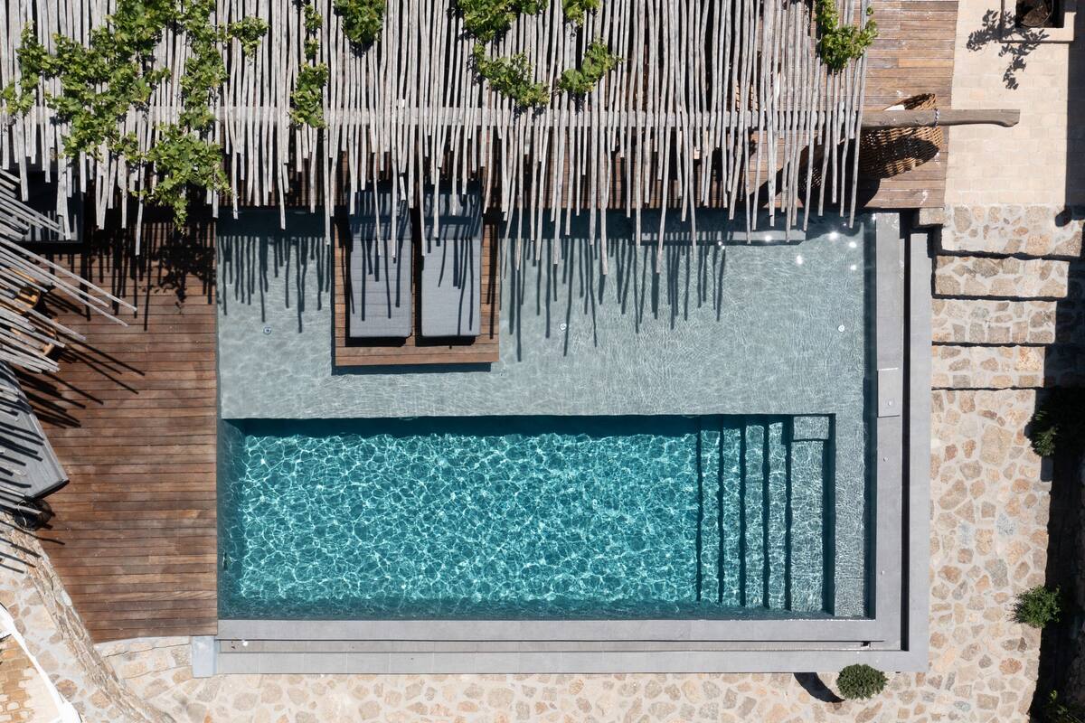 An aerial view of a rectangular swimming pool with a wooden deck and lounge chairs, surrounded by stone walls and greenery.