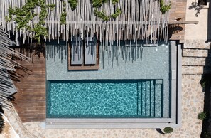 An aerial view of a rectangular swimming pool with a wooden deck and lounge chairs, surrounded by stone walls and greenery. - Ikies of Mykonos (Mykonos)