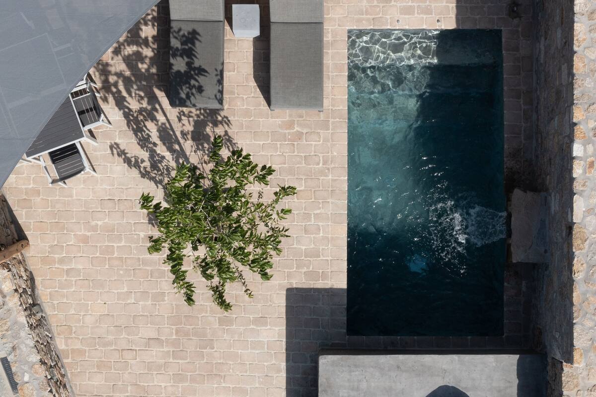 An aerial view of a rooftop pool area with a clear blue pool, stone walls, and a seating area with cushions.