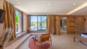 A spacious living area with a large window, a wooden chair, a bench, and a view of a pool and outdoor scenery. - Ikies of Mykonos (Mykonos)