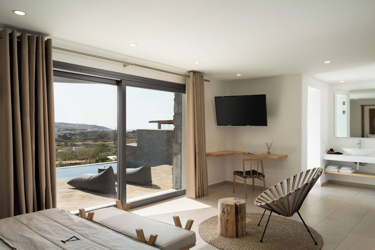 A modern bedroom with a large sliding glass door leading to a balcony, a flat-screen TV mounted on the wall, a wooden side table, and a minimalist bathroom with a large mirror.