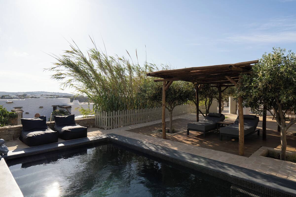 A pool area with lounge chairs and a pergola, surrounded by greenery and overlooking a distant landscape.