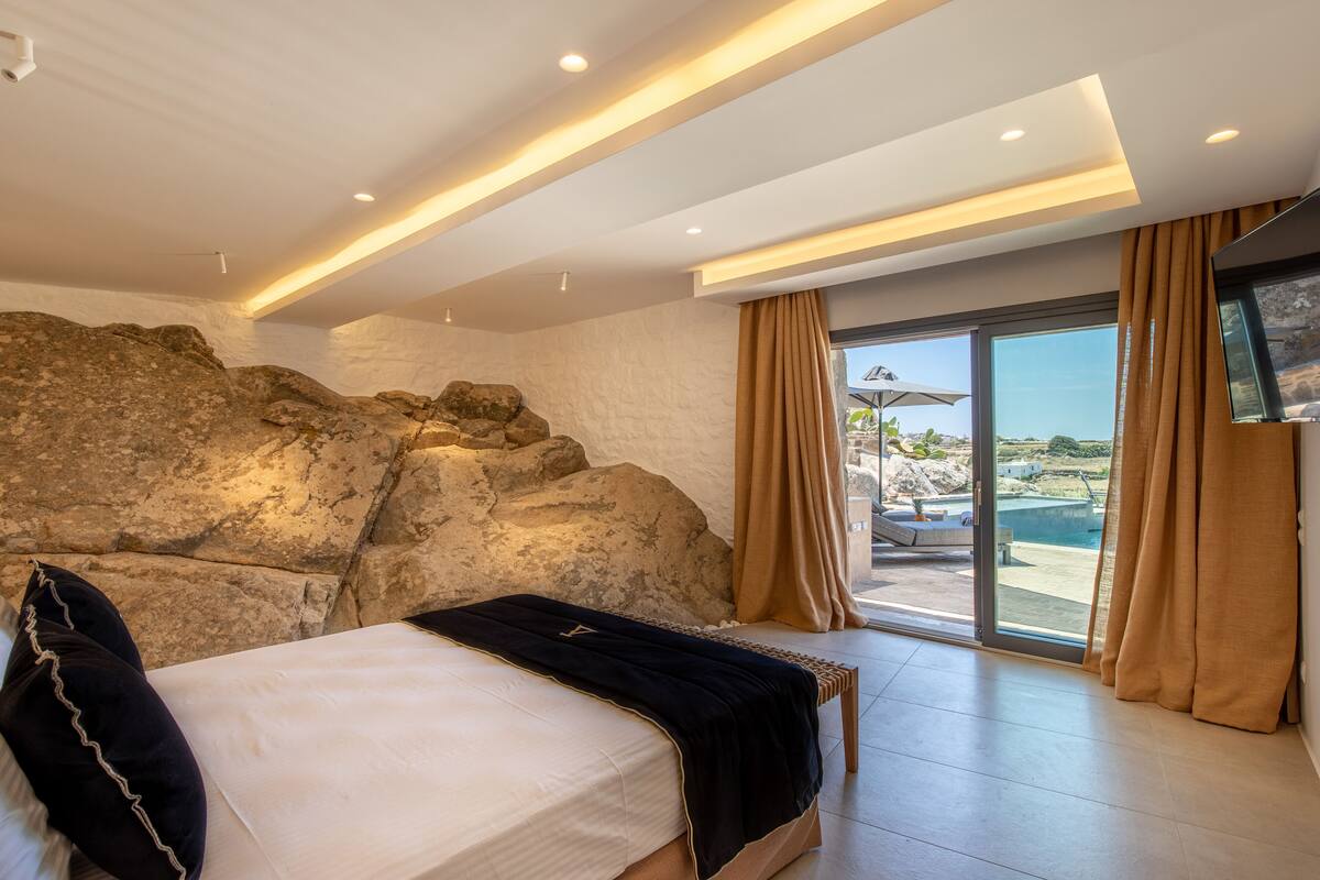 A bedroom with a large bed, a view of the outdoors, and a stone wall.