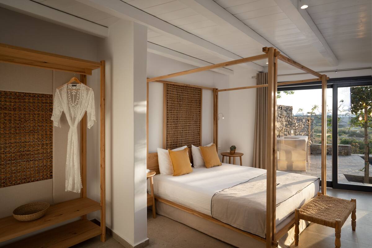 A four-poster bed with a woven headboard, a hanging robe, a small round table, and a wicker stool in a modern bedroom with a view of the outdoors.