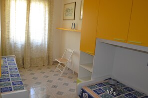 A small, compact room with a bed, a chair, a shelf with items, and a window with curtains.