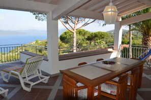 A terrace with a wooden table and chairs, a cushioned chair, and a view of the sea and hills.