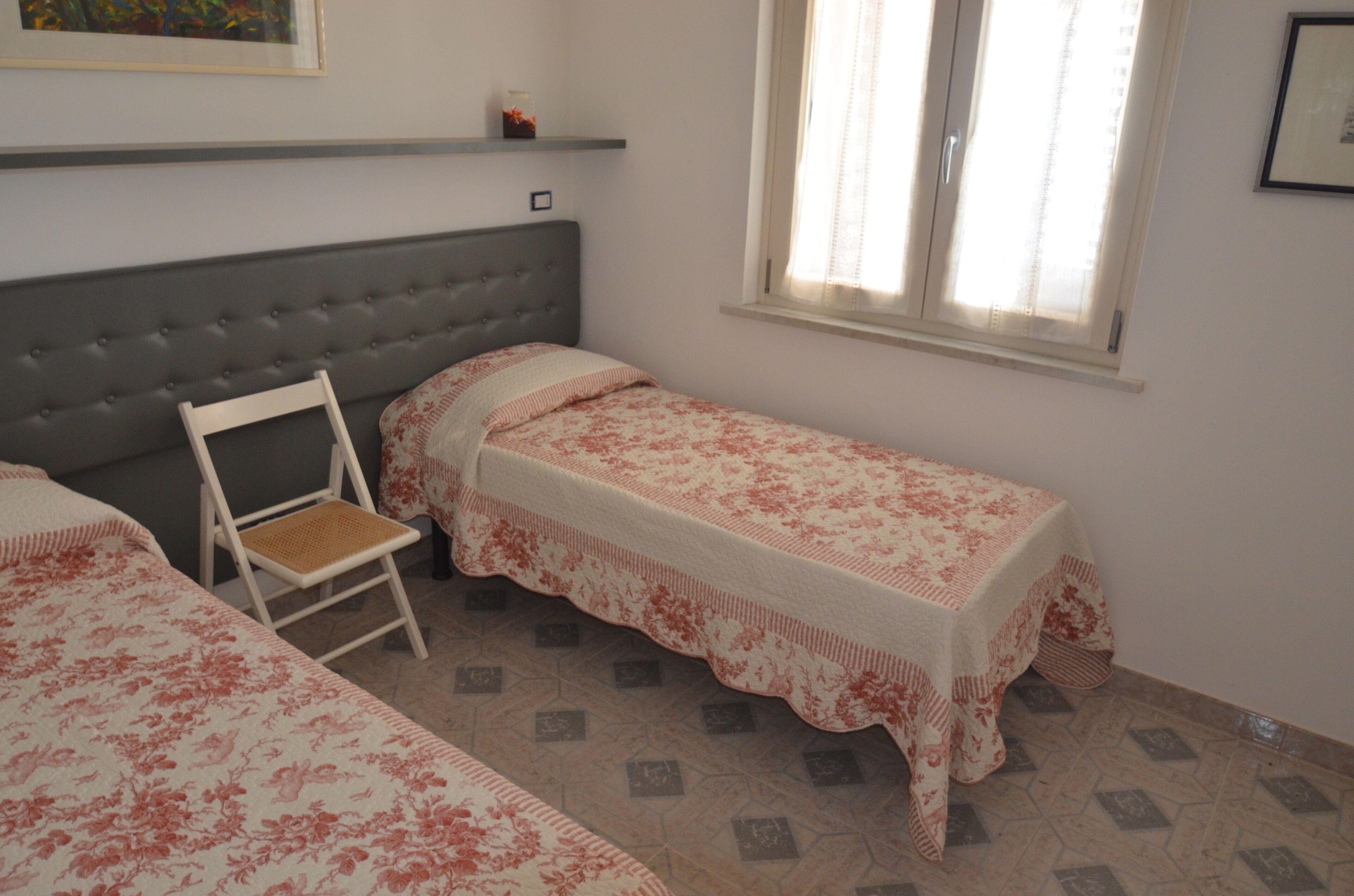 A bedroom with two single beds, a chair, a window with curtains, and a shelf above the bed.