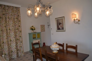 A dining area with a wooden table, chairs, a floral curtain, a green cabinet, a wall-mounted light, and a framed picture.
