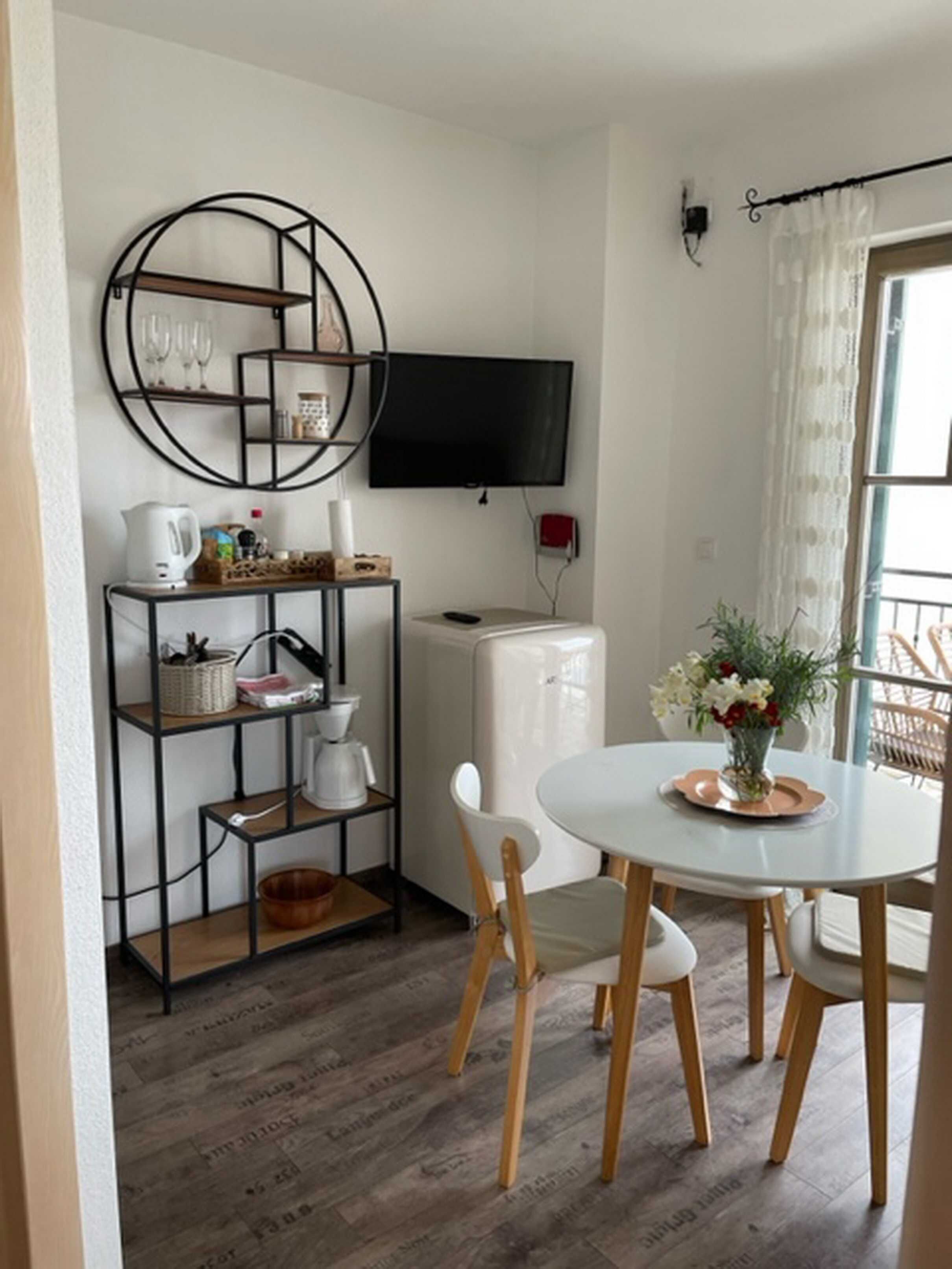 A compact living space with a dining area featuring a round table and two chairs, a kitchenette with a built-in oven, a TV mounted on the wall, and a window with sheer curtains.