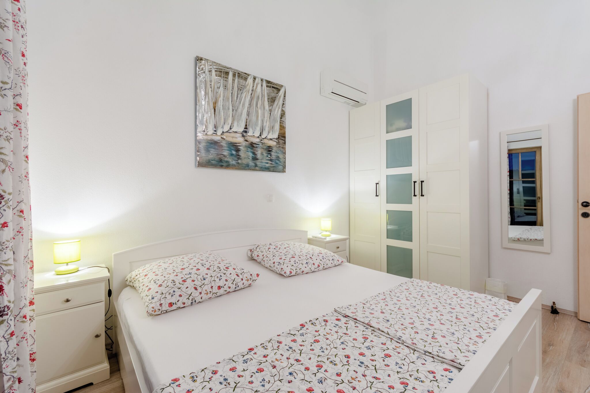A neatly arranged bedroom with a bed, bedside lamps, a nightstand, a wardrobe, and a painting on the wall.