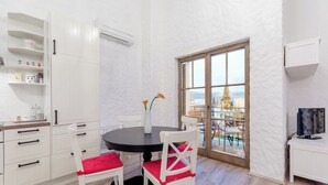 Ein Essbereich mit einem runden Tisch, weißen Stühlen mit roten Kissen, einer Blumenvase und Blick auf einen Balkon mit Gartenmöbeln.