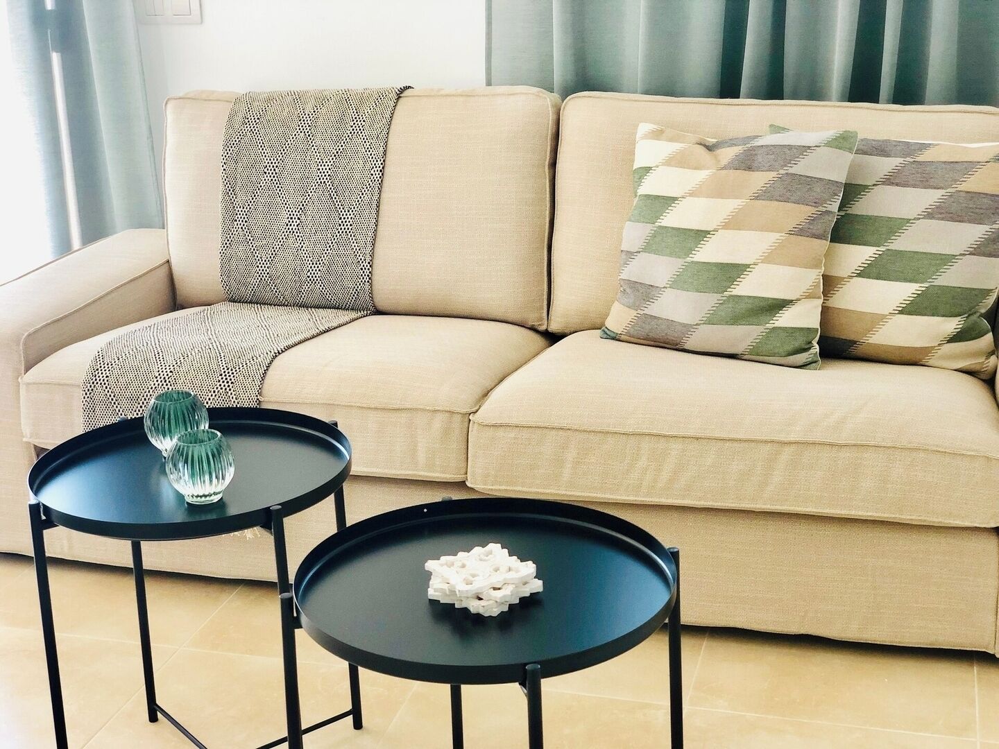 Un canapé beige agrémenté de coussins à motifs, deux tables noires ornées d’objets décoratifs et une fenêtre avec des rideaux.