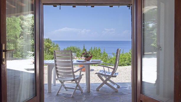 An open door leading to a patio with a table and two chairs, overlooking a scenic view of the sea and greenery. - Villetta dello Scrittore (Capoliveri)