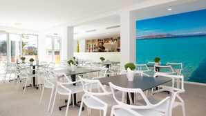 A modern dining area with white chairs and tables, a bar in the background, and a large mural of a coastal scene. - Mix Smart (Llucmajor)