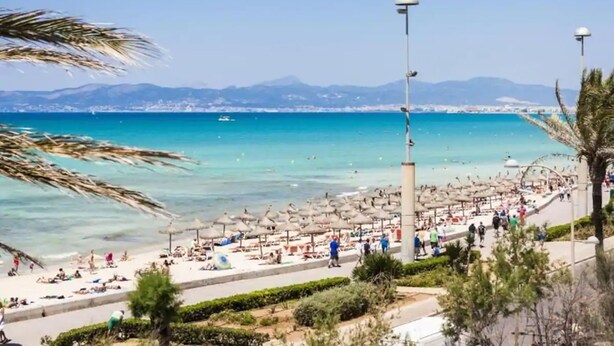 A coastal promenade with beach umbrellas, people, and a clear view of the sea and distant mountains. - Mix Smart (Llucmajor)