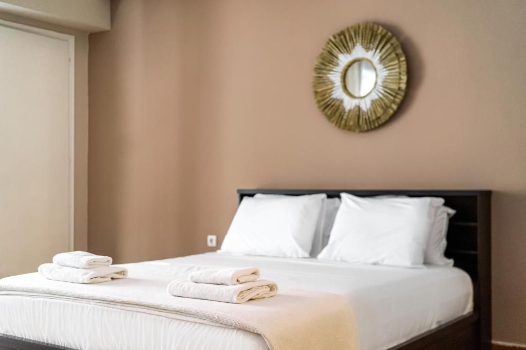 Una cama bien hecha con sábanas blancas y una manta color beige, un espejo con marco dorado en la pared, y una ventana a la izquierda.
