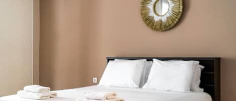 Una cama bien hecha con sábanas blancas y una manta color beige, un espejo con marco dorado en la pared, y una ventana a la izquierda.