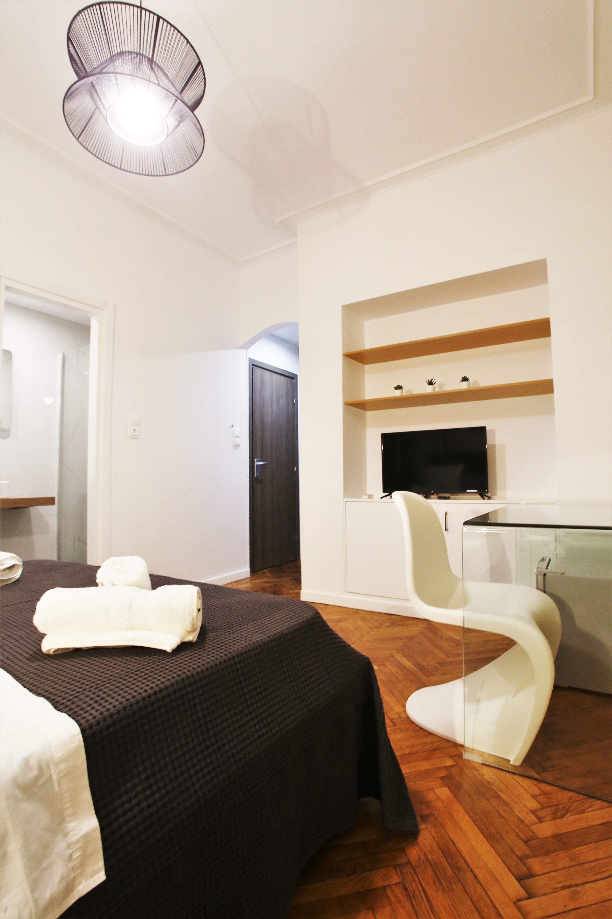 A modern hotel room with a bed, a glass table, a white chair, a TV, and a wooden floor.
