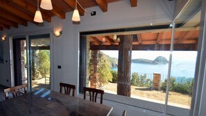 Zona pranzo con un tavolo grande e sedie, ampie finestre vista mare e colline, travi in legno al soffitto.