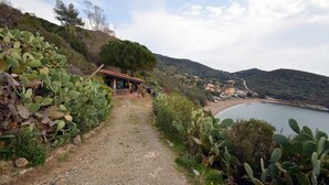 Un sentiero costiero con cactus, un piccolo edificio con tetto rosso e vista sulla spiaggia e le colline sullo sfondo.